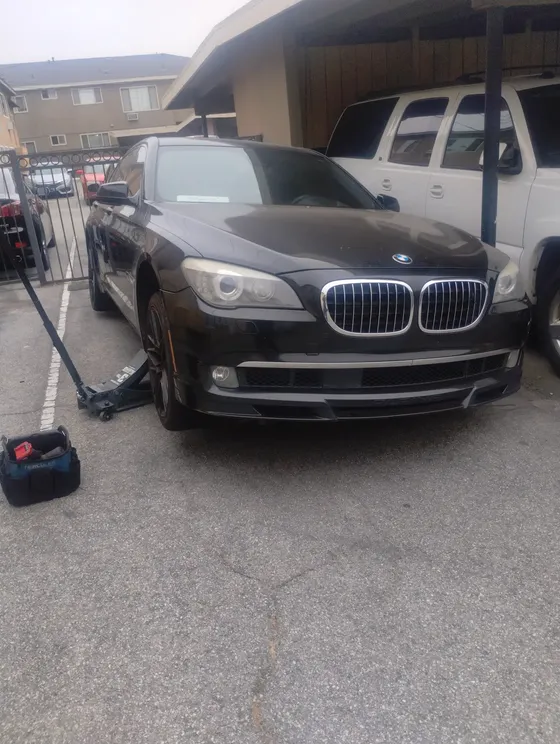 A black sedan raised on a jack at the front in an apartment driveway, a jump pack and tools on the ground beside it.