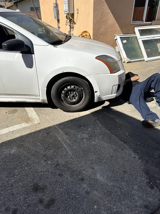 A white sedan on a jack at the curb, a mechanic working underneath the front wheel.
