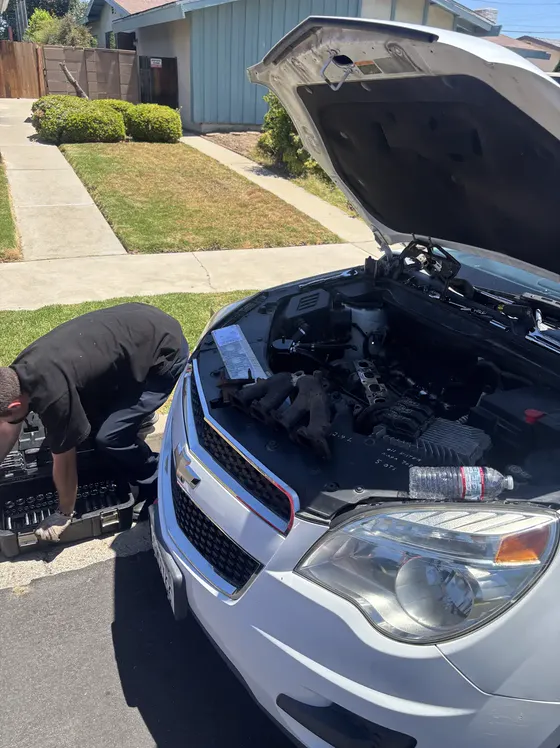 A mechanic kneeling at the front of an SUV with its hood up in a residential driveway.